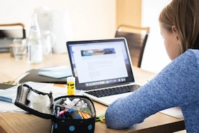 Image of a girl using a Macbook on a table with stationaries and school supplies.