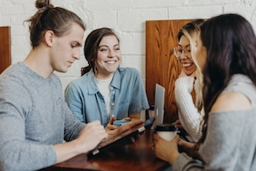 An image of a group of 3 females and 1 male collaborating across a rustic table with a Macbook, coffee, and Tablet/Ipad.