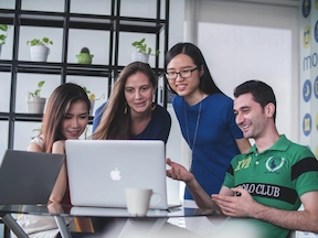 An image of a group 3 females and 1 male collaborating looking at a Macbook