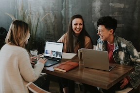 An image of a group of 2 females and 1 male collaborating across a table with a Samsung laptop and a Samsung chromebook, with a leather binded book and drinks.